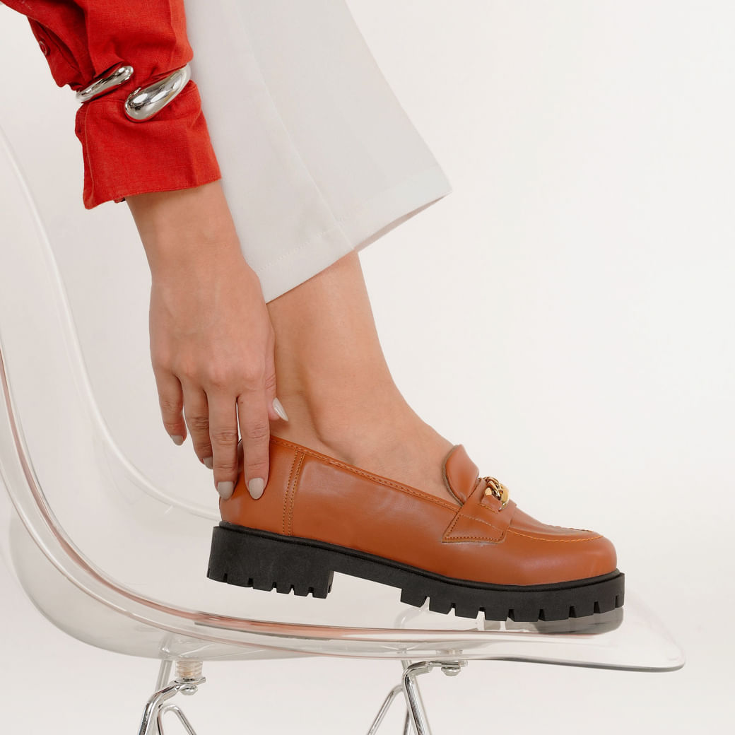 Mocassim feminina M Shuz Whisk Napa Soft marrom vista lateral sobre cadeira transparente com detalhe de corrente dourada e sola tratorada preta.